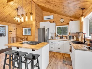 Cocina moderna con detalles de madera, una isla central con taburetes, armarios blancos, electrodomésticos de acero inoxidable e iluminación colgante.