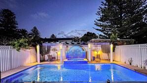 Ein Swimmingpool mit integriertem Whirlpool, umgeben von einem weißen Zaun und einer Pergola mit Beleuchtung.