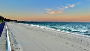 Ein weiter, sandiger Strand mit einer Metallreling auf der linken Seite, die zu einer Stadtansicht in der Ferne führt.