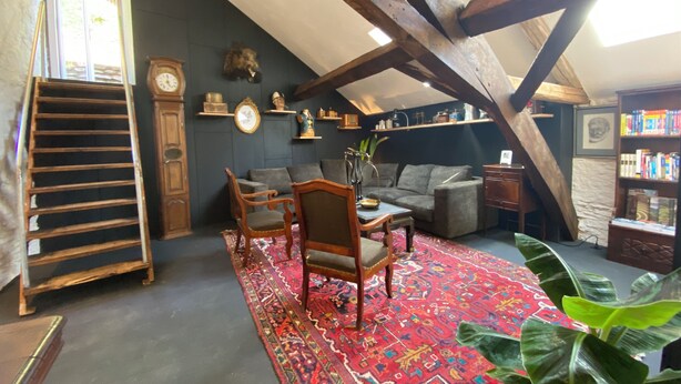 Una habitación acogedora en el ático, con techo inclinado, vigas de madera, un tapete rojo estampado, un sofá, una silla de madera, una mesita auxiliar, una estantería y una planta grande.
