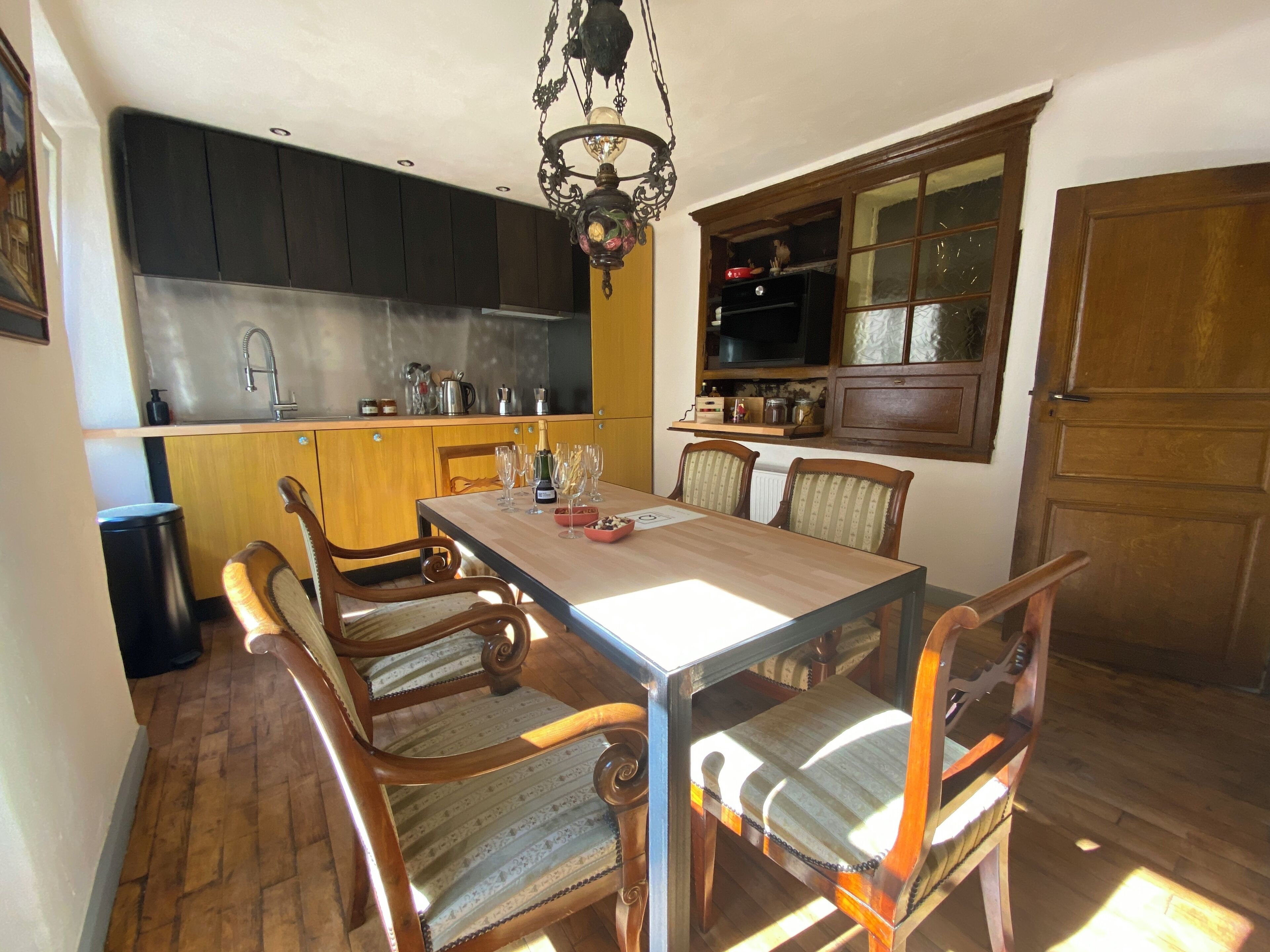 A dining area with a wooden table and chairs, a chandelier, and a kitchen in the background.