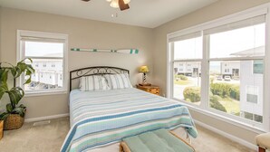A bedroom with a bed, a nightstand, a lamp, a ceiling fan, and large windows offering a view of the neighborhood. - Shangri La - 2BR Turtle Cove Townhouse Near Pool (Surf City)