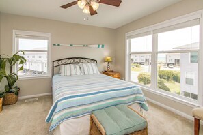 Ein Schlafzimmer mit Bett, Nachttisch, Lampe, Deckenventilator und großen Fenstern mit Blick auf Häuser.