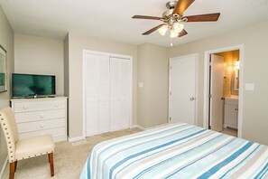 Ein Schlafzimmer mit einem Bett, einem Sessel, einem Fernseher auf einer Kommode und einem Deckenventilator.