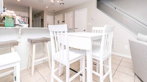 A modern kitchen with white cabinetry, a dining area with white chairs, and a staircase leading to another level. - Shangri La - 2BR Turtle Cove Townhouse Near Pool (Surf City)