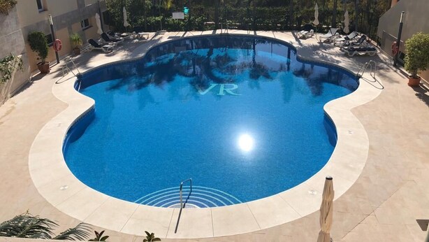 Una piscina con borde ondulado, rodeada de sillones de descanso y sombrillas, con edificios y árboles al fondo.
