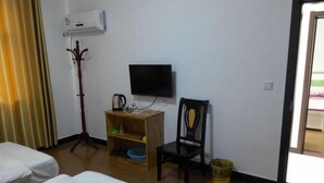 Habitación de hotel con cama, televisor montado en la pared, mesita de madera con tetera, una silla y un estante pequeño con un recipiente azul.