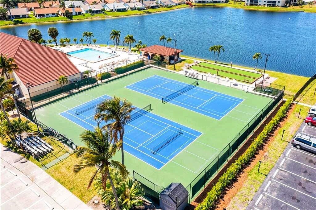 A tennis court with a pool adjacent to it, surrounded by palm trees and a lake.