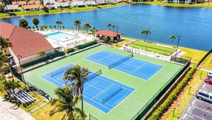 A tennis court with a pool adjacent to it, surrounded by palm trees and a lake. - First floor Cinnamon Cove Condo - Close to SWFL Beaches! (Fort Myers)