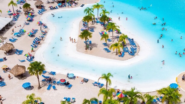 A beach with multiple sun loungers and umbrellas, a central pool area, and palm trees. - First floor Cinnamon Cove Condo - Close to SWFL Beaches! (Fort Myers)