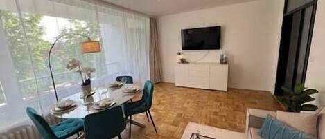 A modern living room with a dining area, a flat-screen TV mounted on the wall, and a large window with curtains.