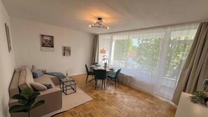 Un salon avec un canapé, une table à manger entourée de chaises, une plante en pot et une grande fenêtre avec des rideaux.
