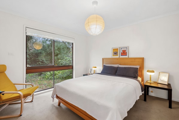 A bedroom with a large window, a bed with a wooden headboard, a bedside table with a lamp, and a chair.