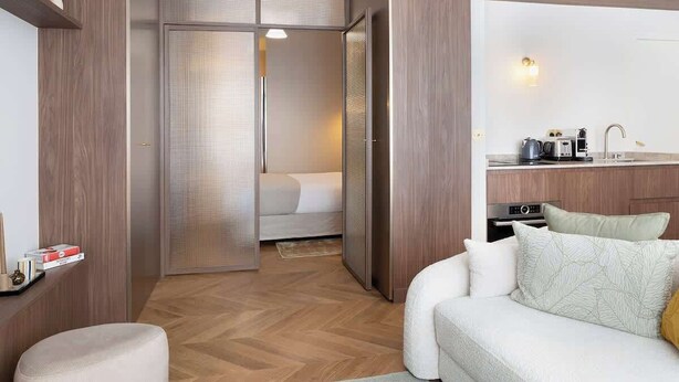 Une chambre d’hôtel moderne avec un plancher en bois, un canapé blanc, un lit avec une couette blanche, une kitchenette équipée d’un évier et d’une cuisinière, et un placard avec une porte coulissante.