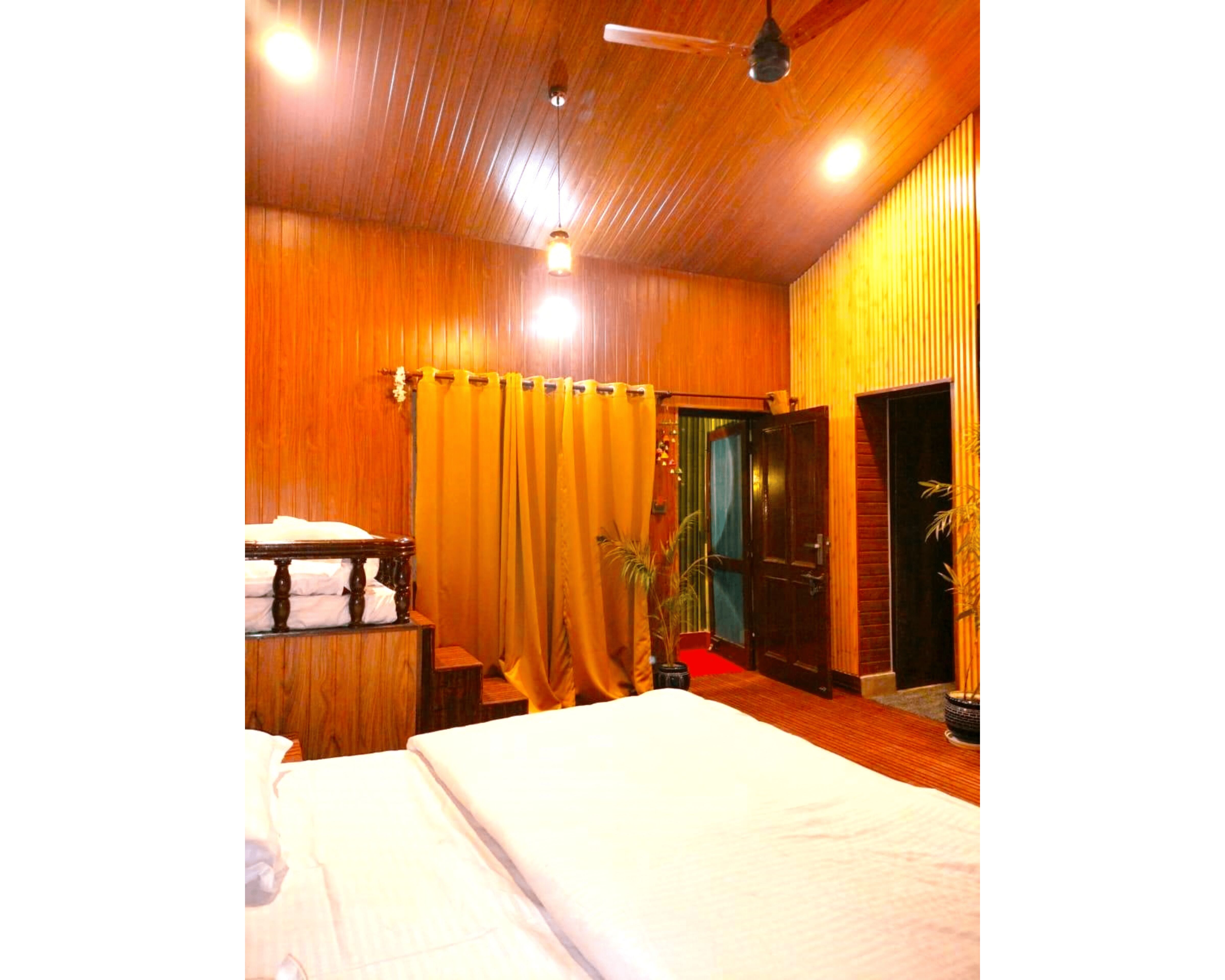Una habitación con paneles de madera, una cama, una mesita de noche, una planta en maceta y una puerta que da a otra habitación.