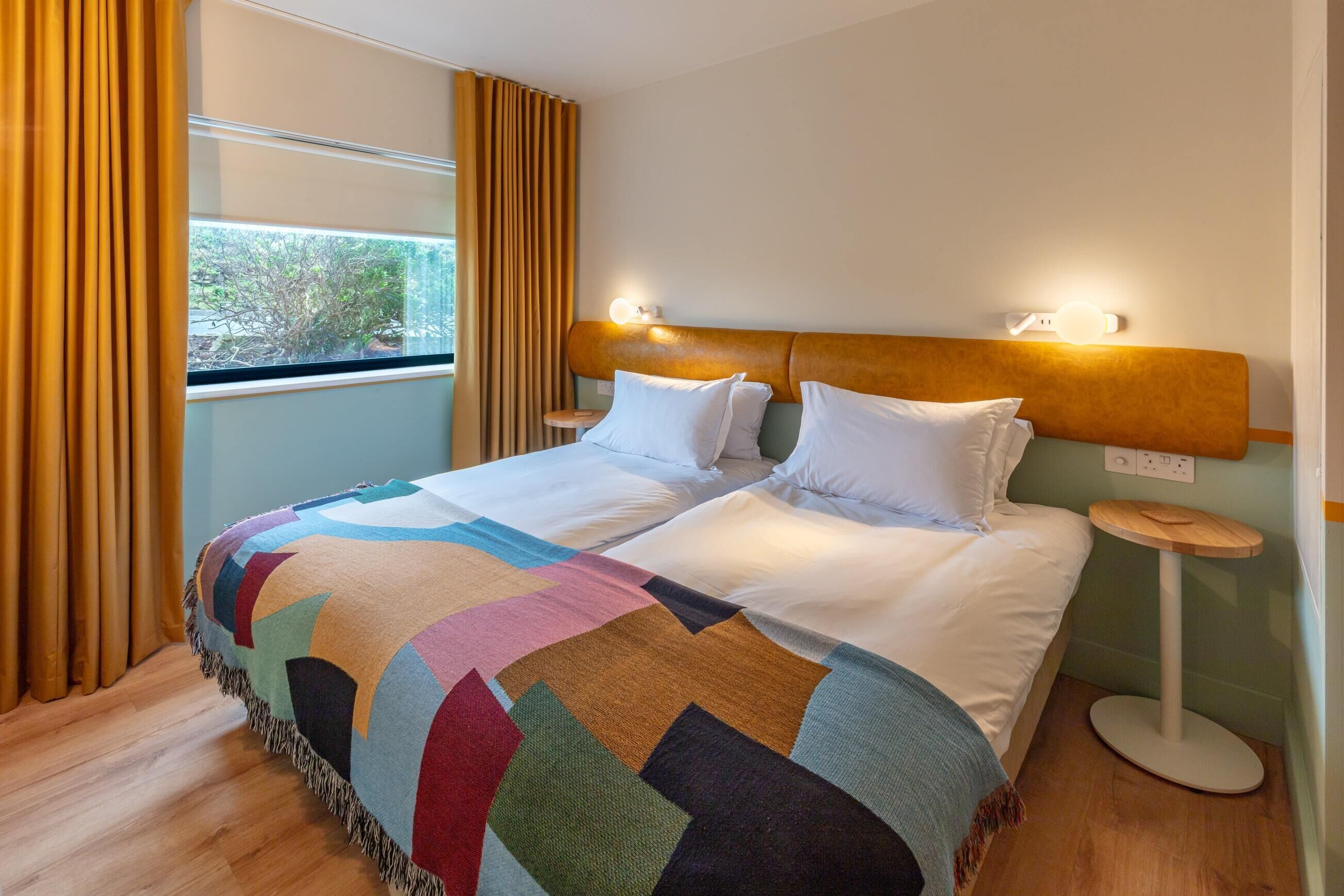 A neatly made bed with a colorful blanket, a headboard with wall-mounted lights, and a bedside table with a lamp.