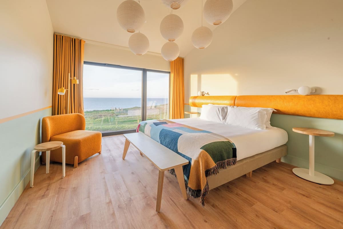 Une chambre d’hôtel moderne avec un grand lit, une banquette, une petite table et une vue sur la mer.