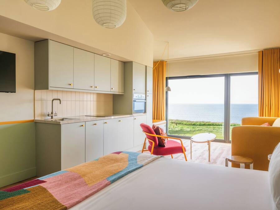 A hotel room with a bed, a small desk, a chair, a TV, and a view of the sea.
