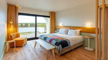 Una habitación de hotel moderna con una cama grande, un banco, una silla, una mesita y vistas al exterior a través de una puerta corrediza.