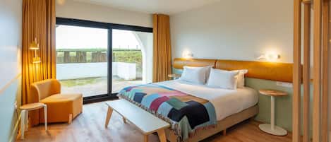 A modern hotel room with a large bed, a bench, a chair, a small table, and a view of the outdoors through a sliding door.