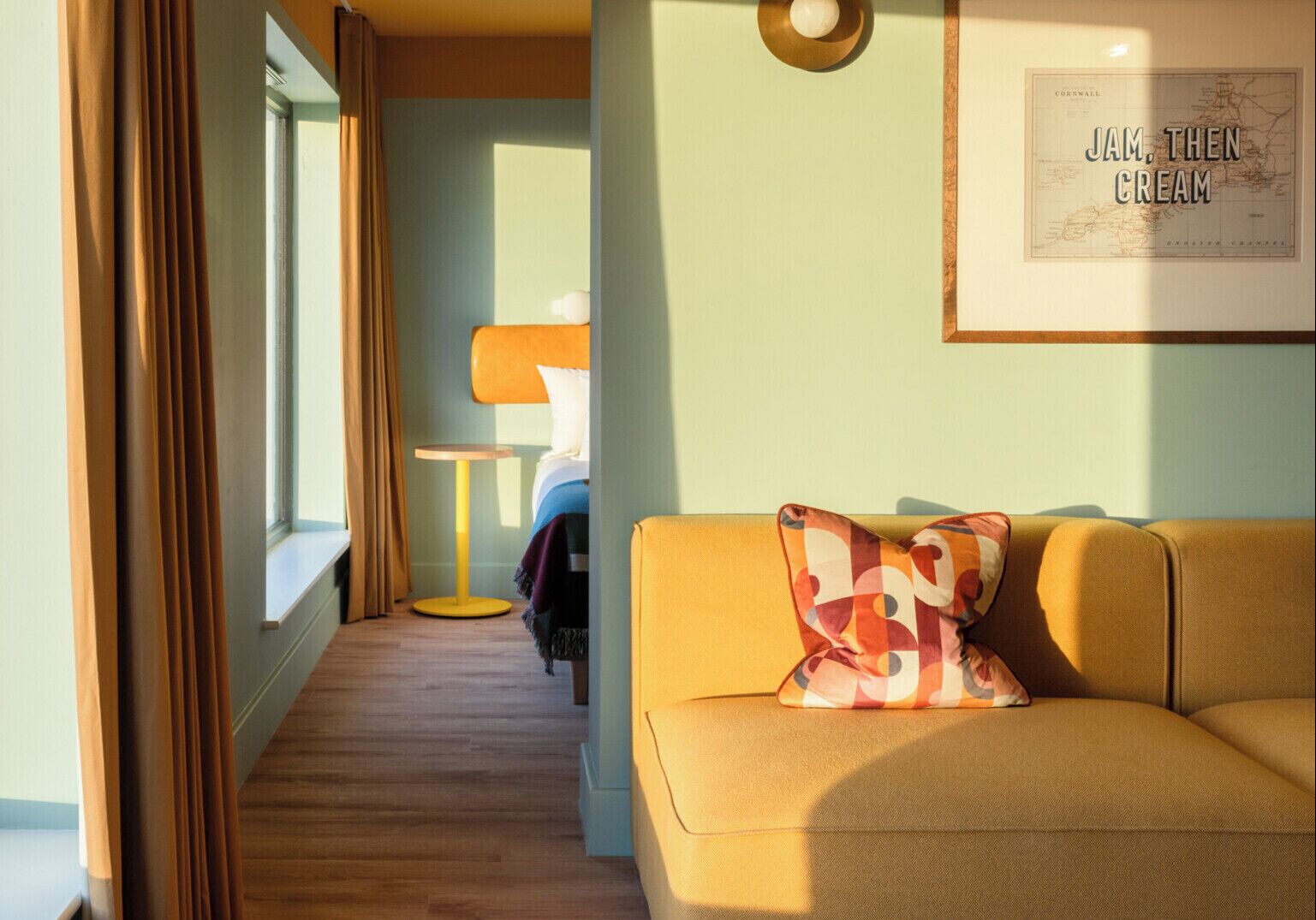Ein Hotelzimmer mit gelbem Sofa, einem kleinen runden Tisch, einem Bett mit weißem Kopfteil und einer gerahmten Karte an der Wand.