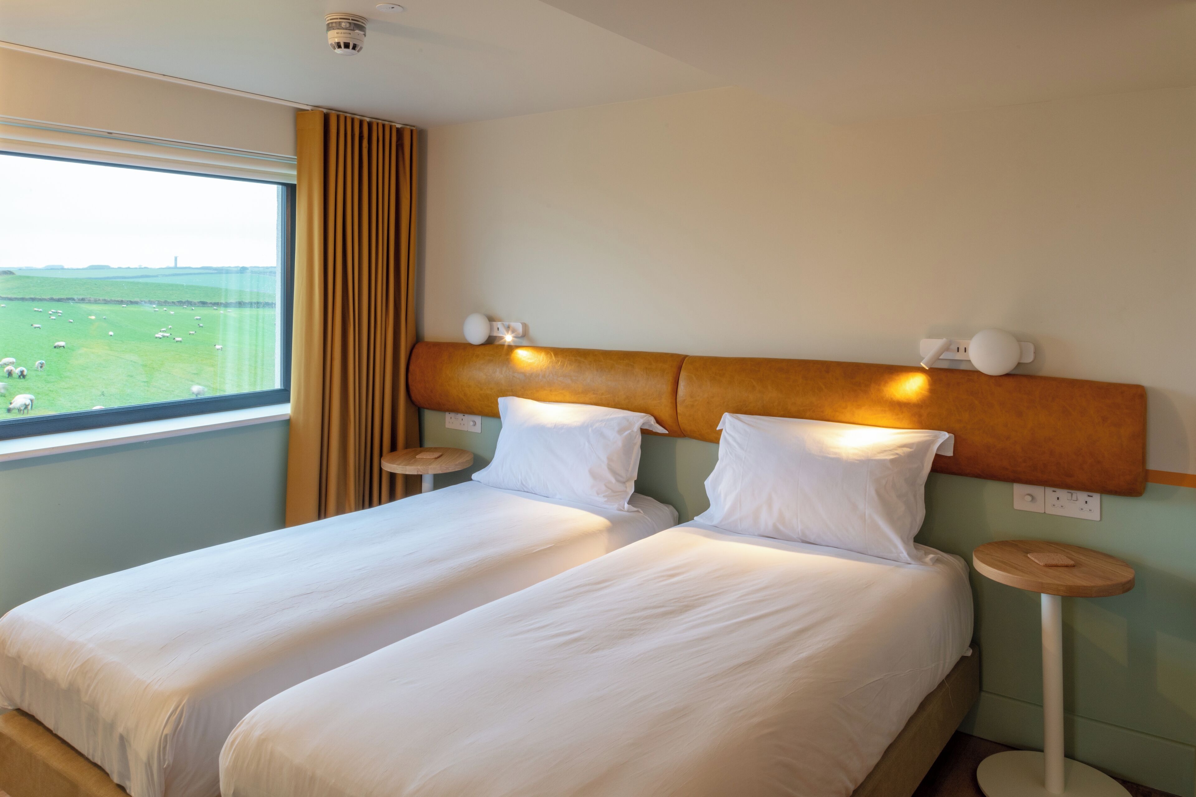 Une chambre d’hôtel avec deux lits, une grande fenêtre donnant sur un champ verdoyant et une tête de lit agrémentée de lampes murales.