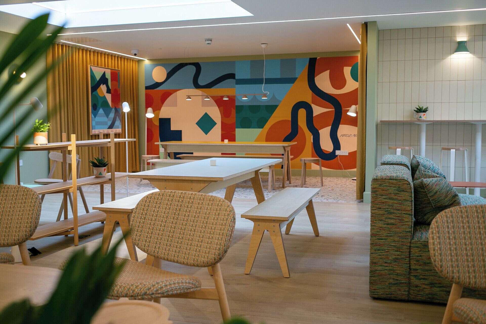 A modern lounge area with wooden tables and chairs, a colorful abstract mural, and a seating area with patterned cushions.