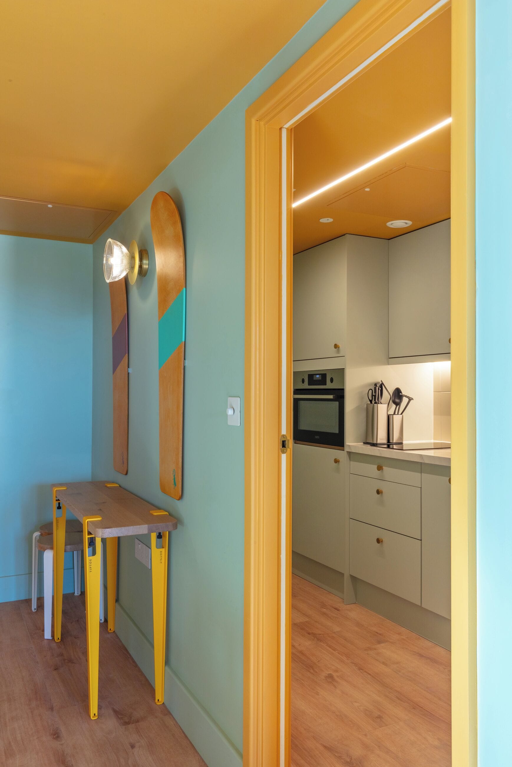 A room with a wooden table and chairs, a wall-mounted light fixture, and a kitchen area with cabinets and a countertop.