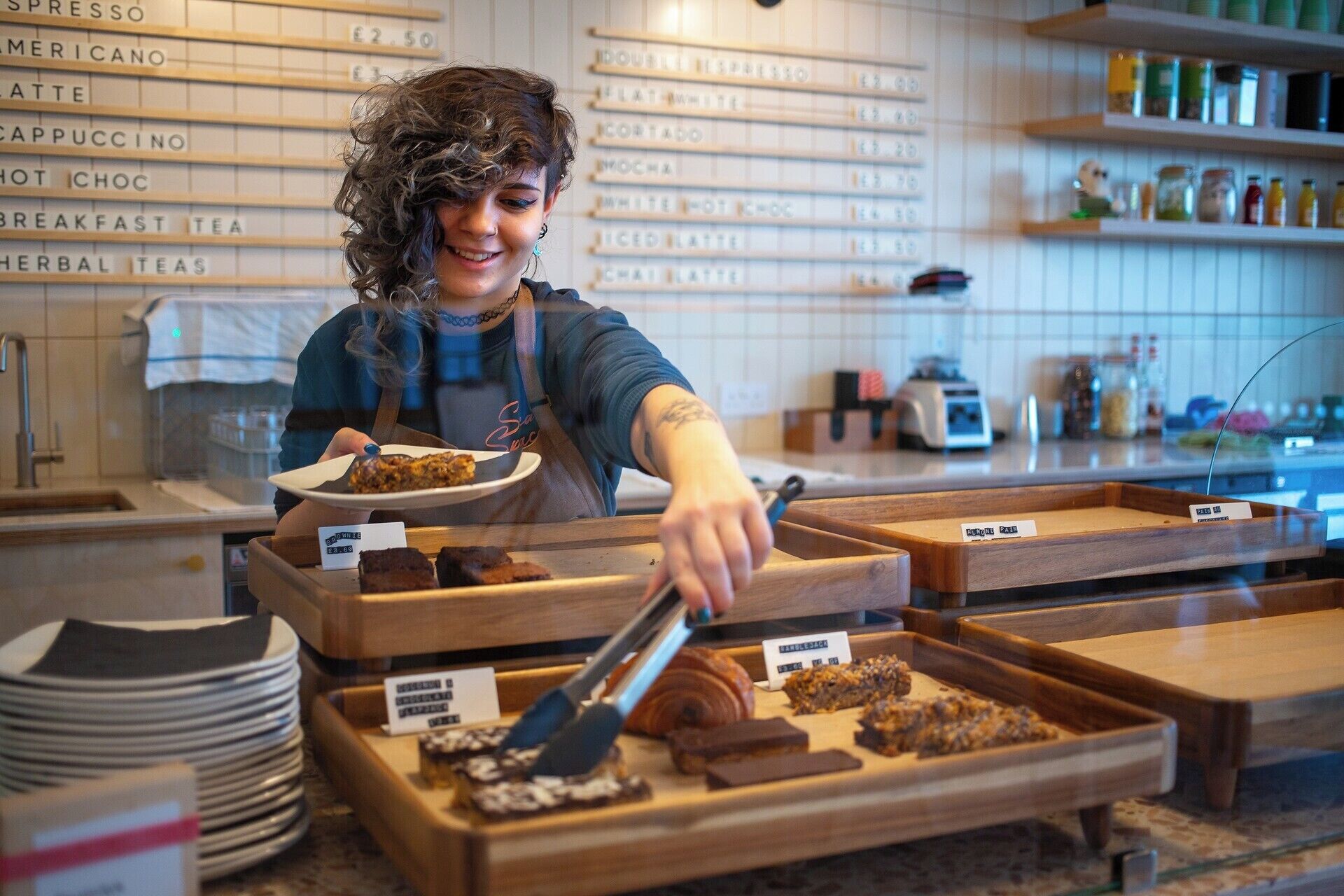 A person serving food in a cafe with wooden tables and a menu board in the background.