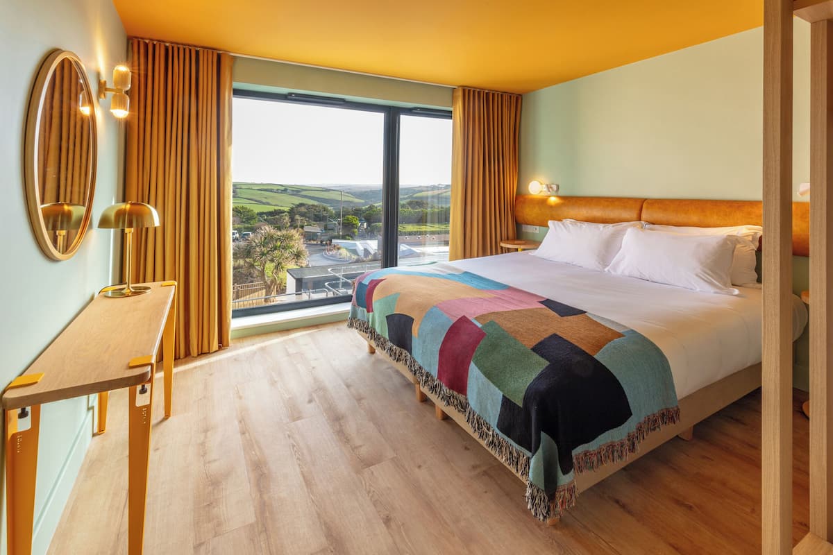 Une chambre d’hôtel avec un grand lit, une table de chevet en bois, un miroir et une vue sur la campagne par une grande fenêtre.