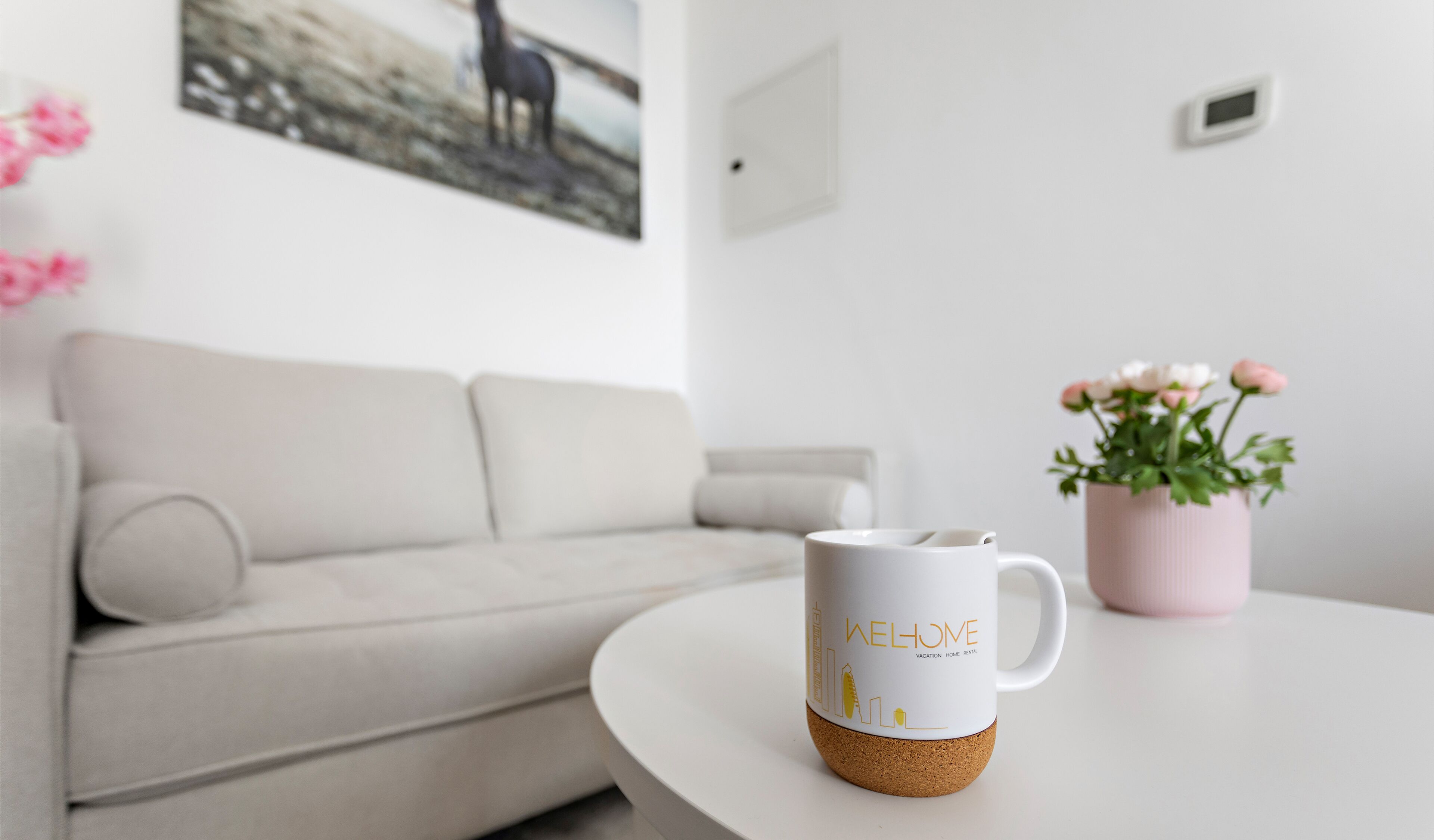 A white mug with text on a white table, a white sofa, and a pink vase with flowers.