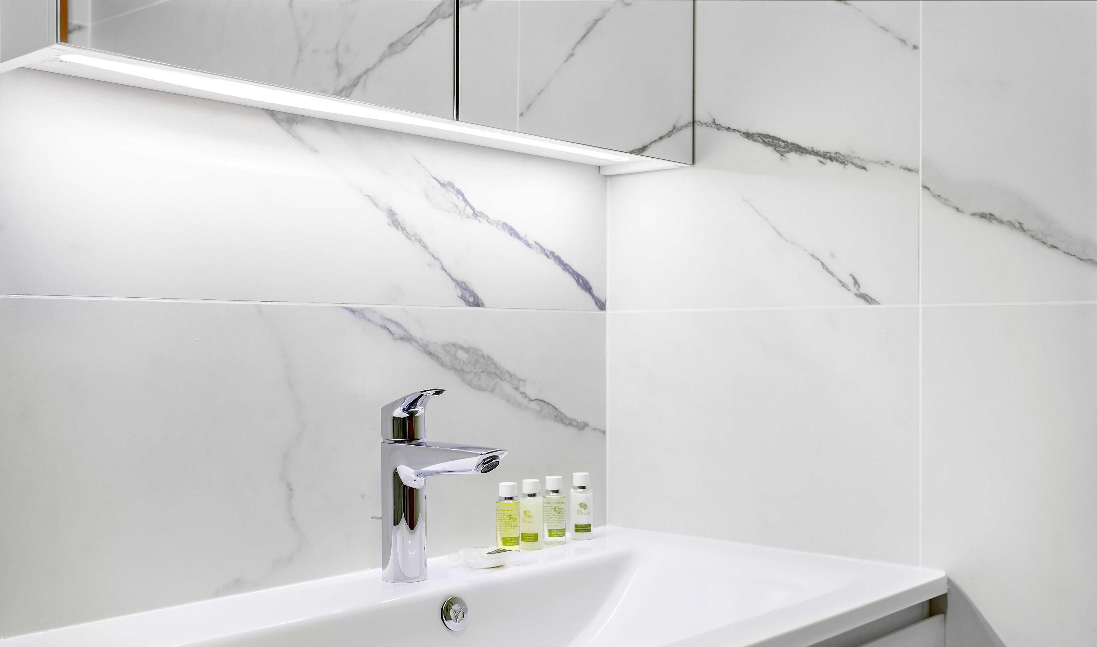 A modern bathroom with a white marble sink, chrome faucet, and a mirror above. There are four bottles of toiletries on the sink.