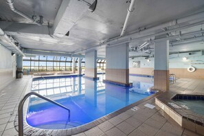 Une grande piscine intérieure à l’eau d’un bleu limpide, avec des murs carrelés et une vue sur le ciel grâce à de grandes fenêtres.