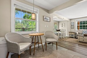 Un coin repas avec des chaises en osier, une table ronde en bois et une grande fenêtre équipée de stores.