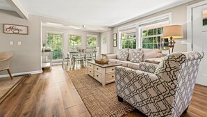 Ein geräumiges Wohnzimmer mit einer Couch, einem Couchtisch und einer Lampe. Im Hintergrund ist ein Essbereich mit Stühlen und einem Tisch zu sehen.