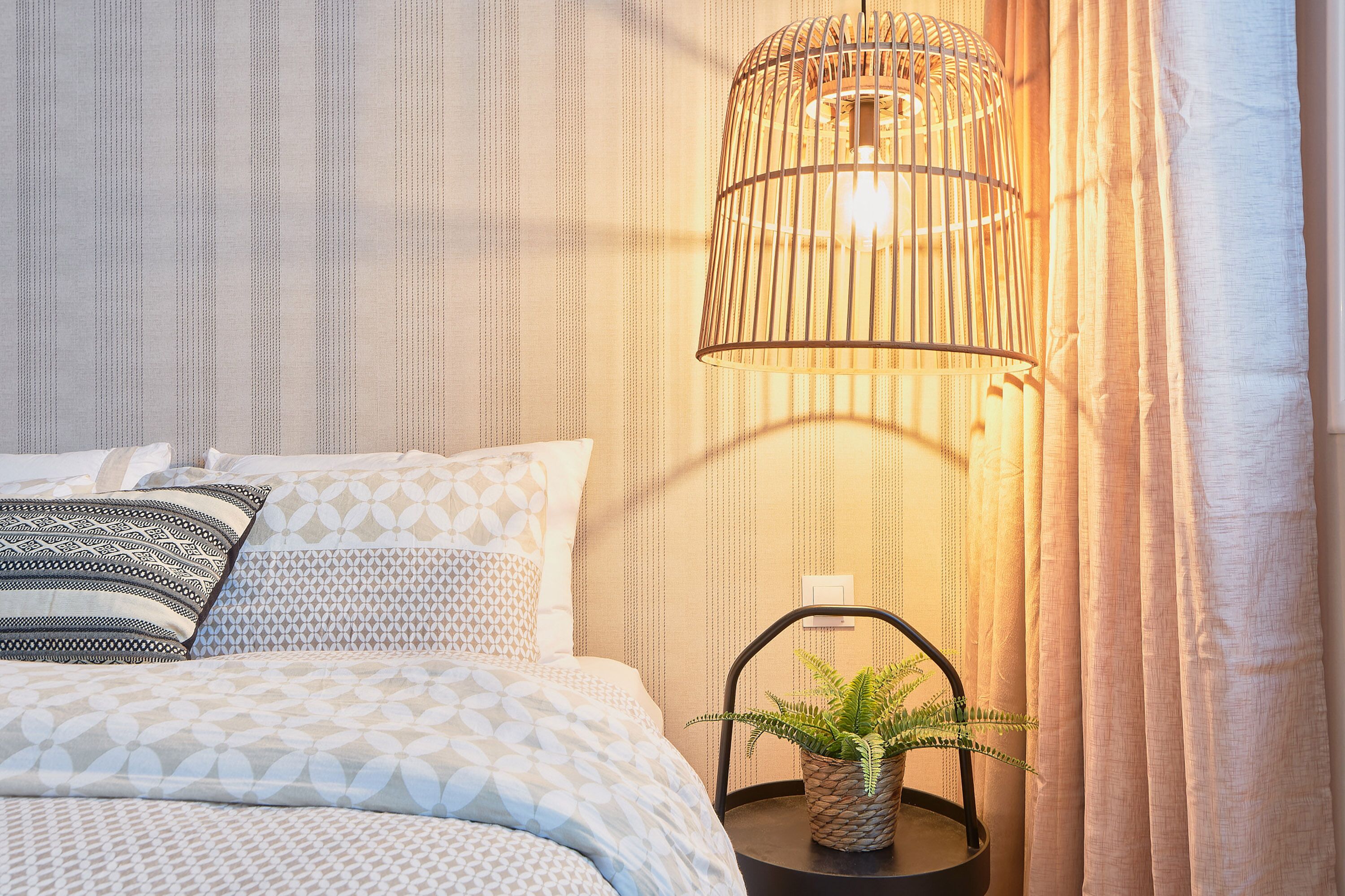 A neatly made bed with patterned bedding, a hanging light fixture, and a potted plant on a bedside table.