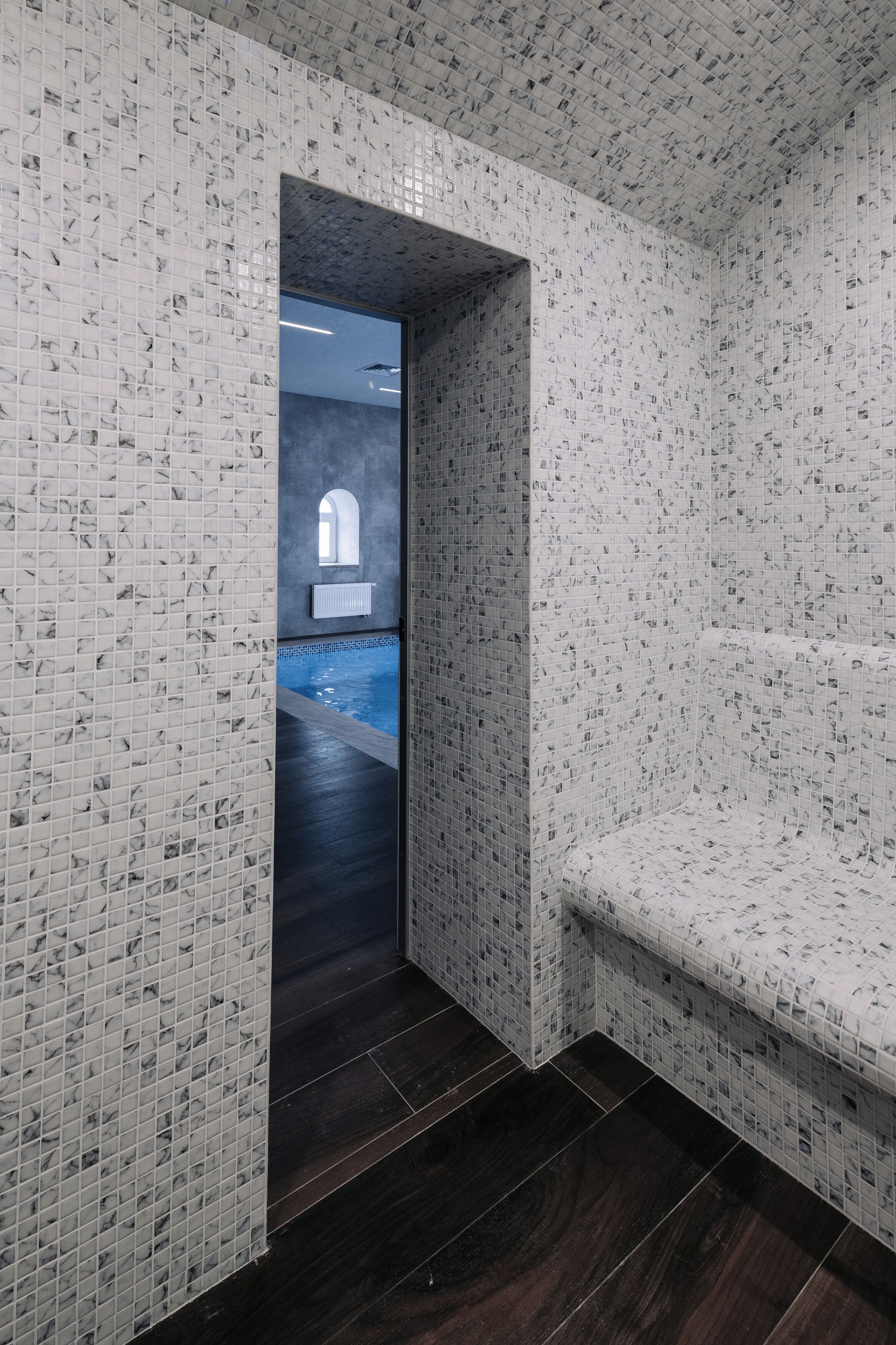 A hallway with mosaic tiled walls and a dark wooden floor, leading to a pool area.