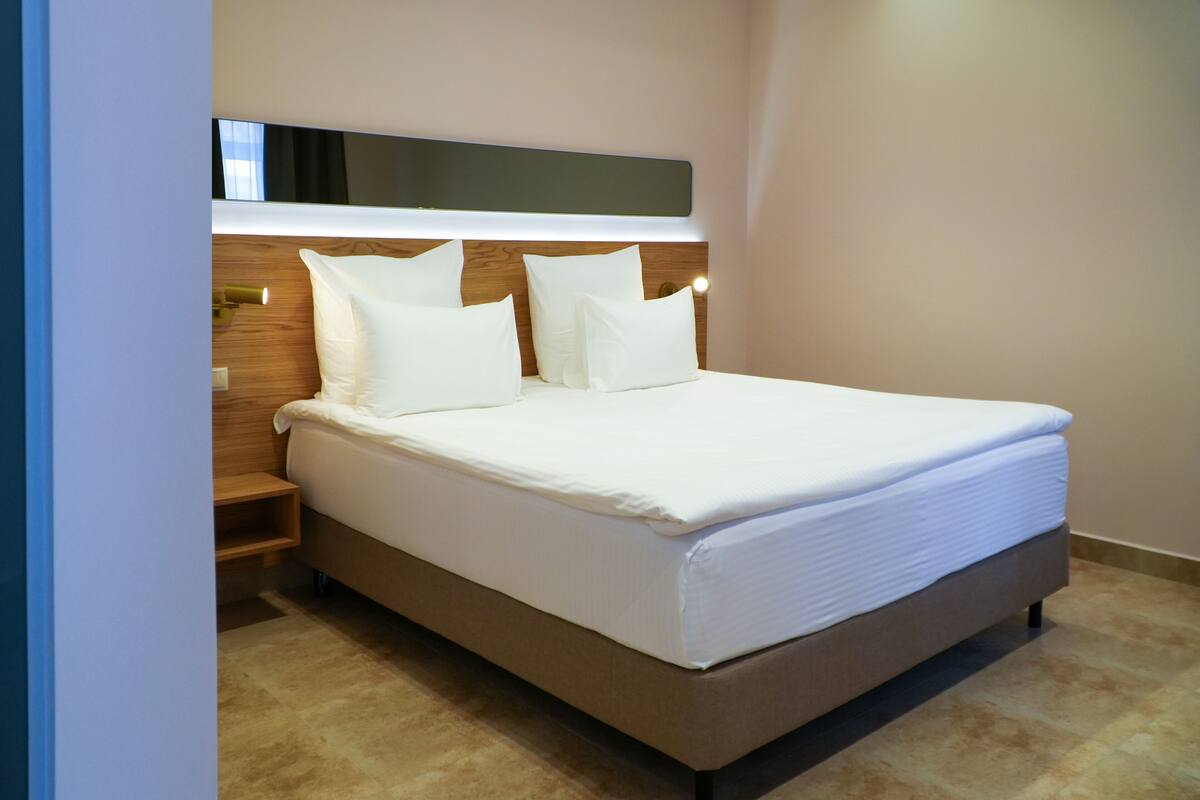 Cama bem arrumada com lençóis e almofadas brancos, cabeceira de madeira e mesa de cabeceira com abajur.