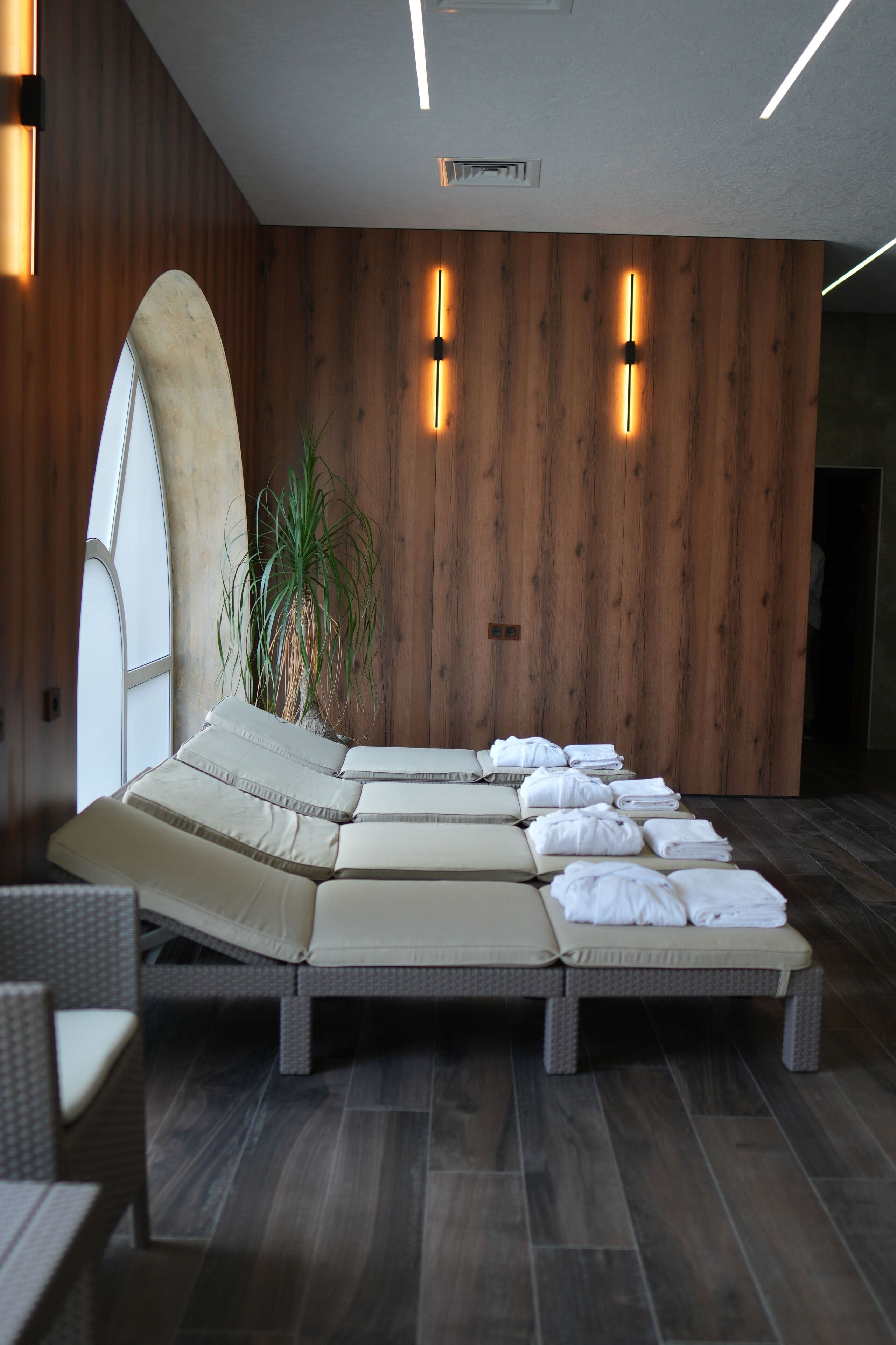 Un'elegante sala spa moderna con un'ampia finestra ad arco, un angolo relax con cuscini, una pianta in vaso e pannelli in legno.
