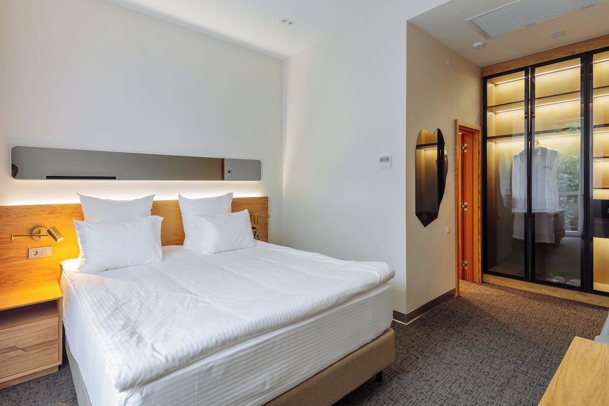 Quarto de hotel moderno com uma cama grande, mesa de cabeceira, guarda-roupa e vista através de uma porta de vidro.