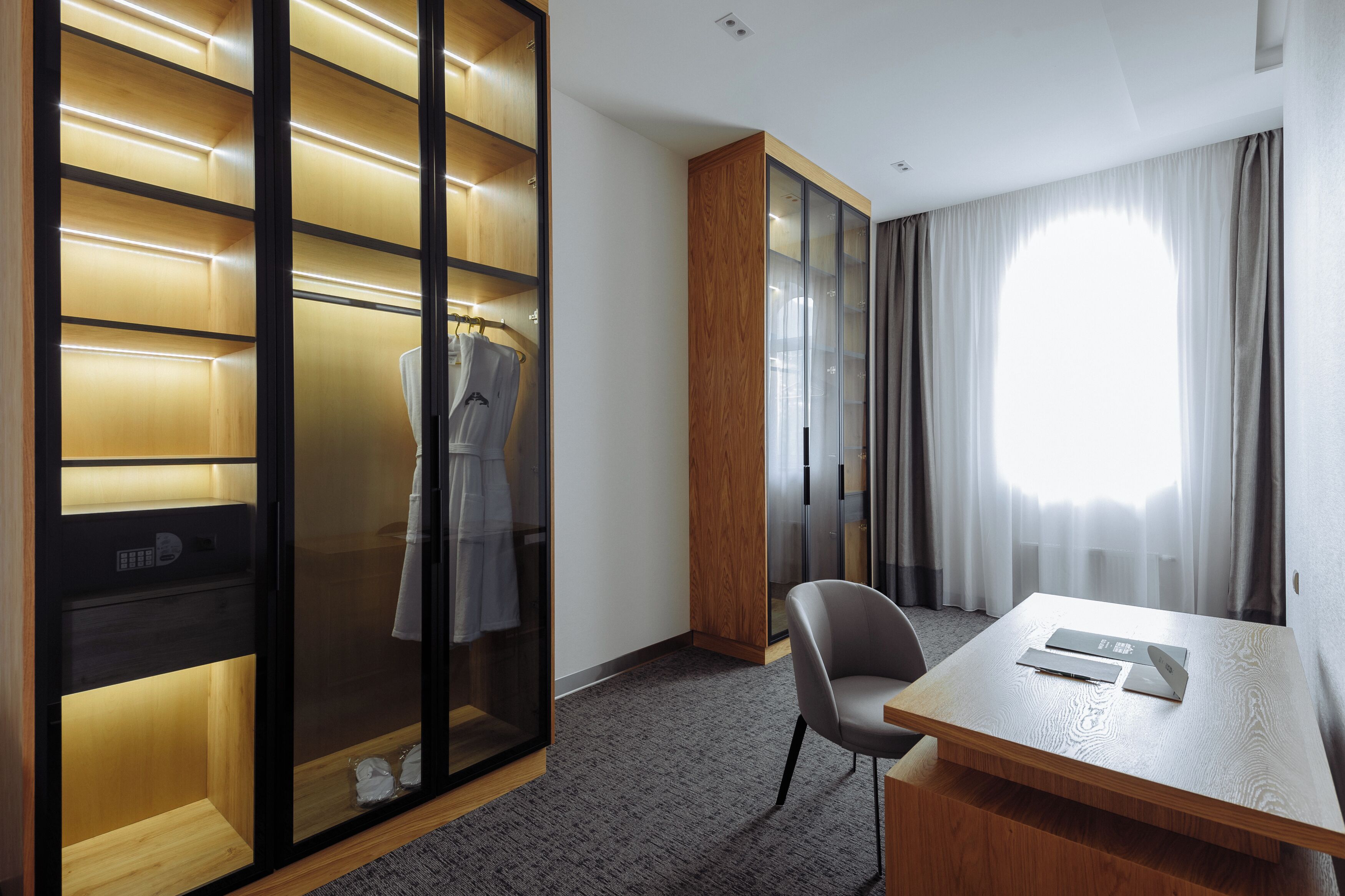 A hotel room with a wooden wardrobe, a desk with a laptop, and a large window with curtains.