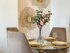 A dining table with a glass top, surrounded by cushioned chairs, set with plates, cutlery, and a vase with flowers.