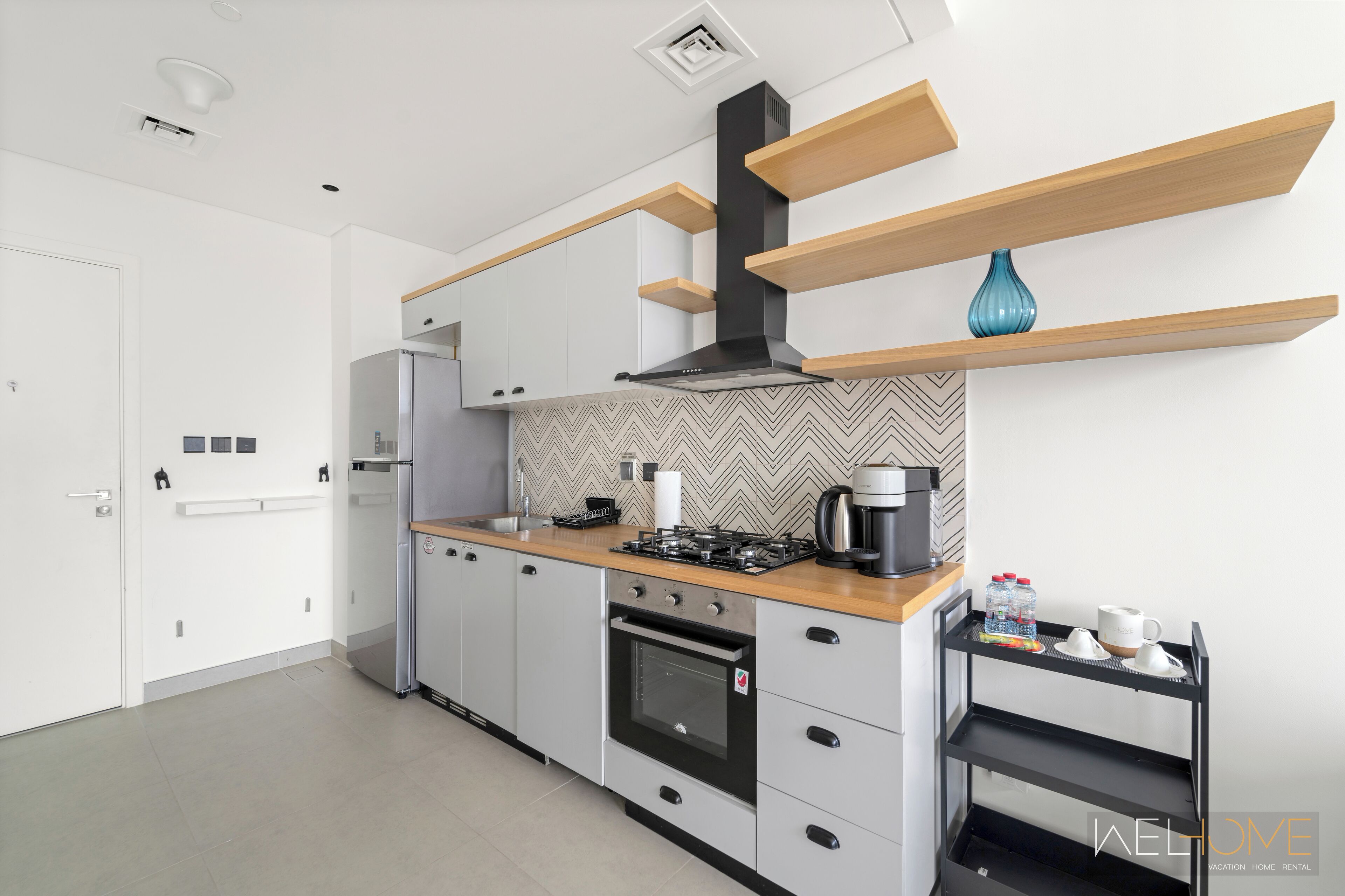 A modern kitchen with grey cabinets, a black stove, and a coffee machine. There are wooden shelves above the counter and a small table with cups and bottles.