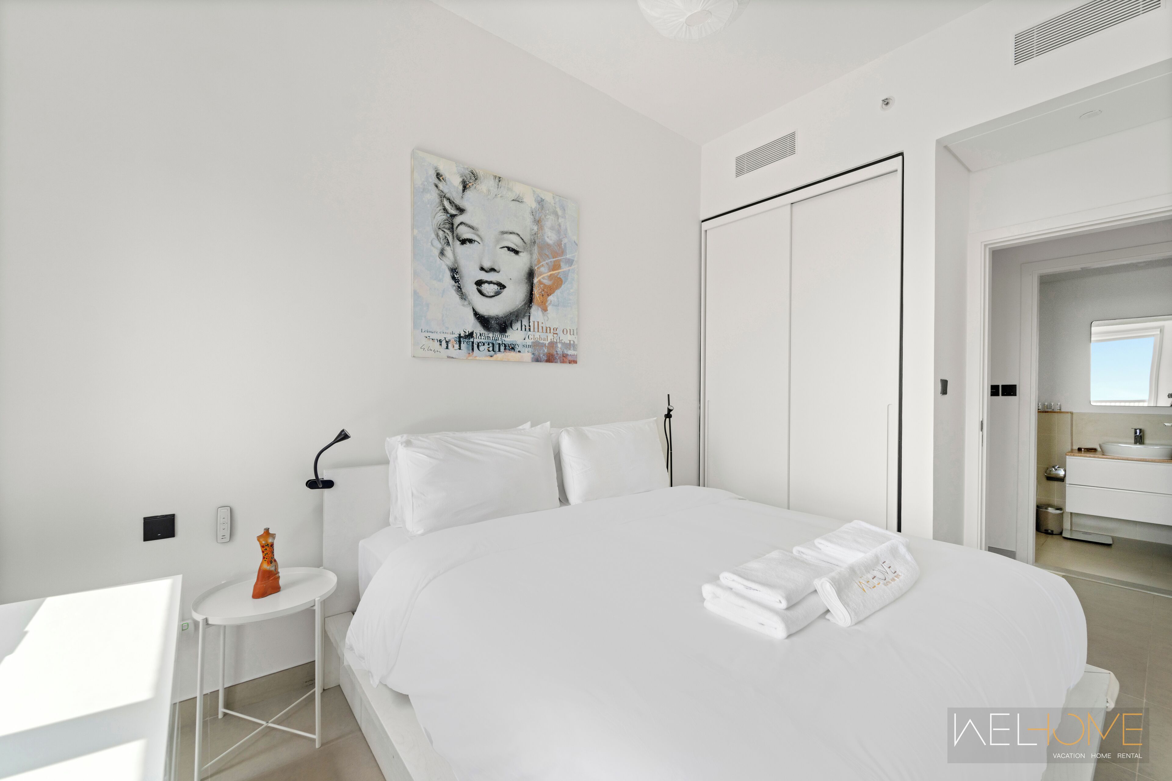 Une chambre d’hôtel moderne avec un grand lit, une petite table ronde, une lampe fixée au mur et une salle de bain visible à travers une porte ouverte.