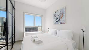 A hotel room with a large bed, a window with a view of buildings, and a painting on the wall. - WelHome - Beautiful 1BR Apt in Dubai Hills with City View (Dubai)