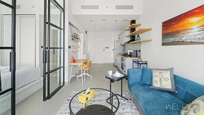 A modern apartment with a glass door leading to a bedroom, a living area with a blue sofa, a kitchen with wooden shelves, and a large painting of a sunset. - WelHome - Beautiful 1BR Apt in Dubai Hills with City View (Dubai)