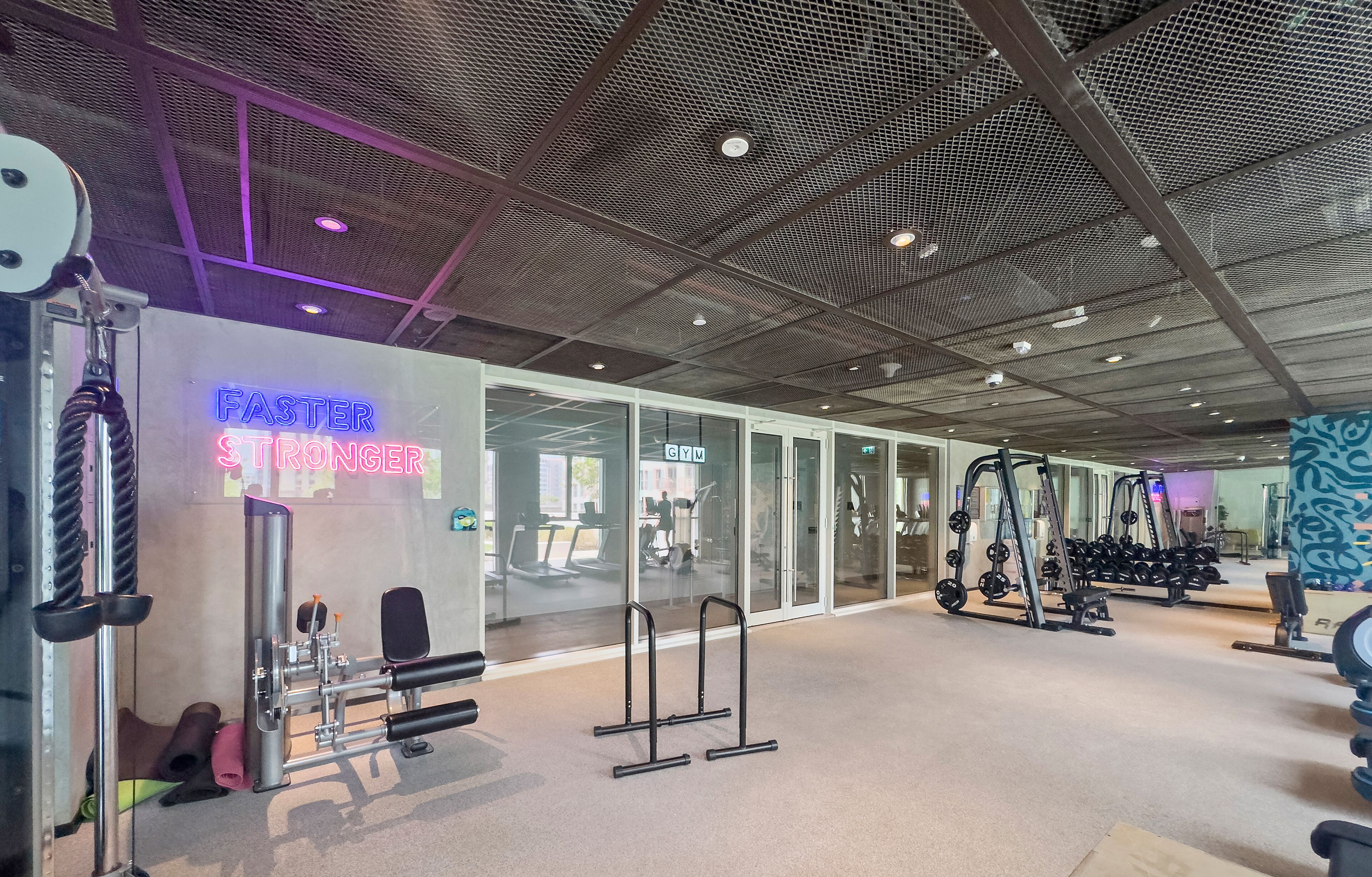 Une salle de sport équipée de divers appareils d’exercice, un panneau lumineux au néon et un plafond orné d’un motif en réseau.
