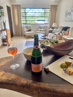 A bottle of Veuve Clicquot rosé champagne, a glass of rosé wine, a bowl of fruit, and a cutting board with fruit on a wooden table in a living room.