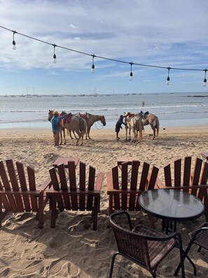 Een zithoek aan het strand met houten stoelen en een ronde tafel, met uitzicht op de oceaan en boten in de verte.