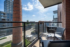 Un balcon avec des fauteuils et une table en osier, donnant sur un parking et des immeubles.
